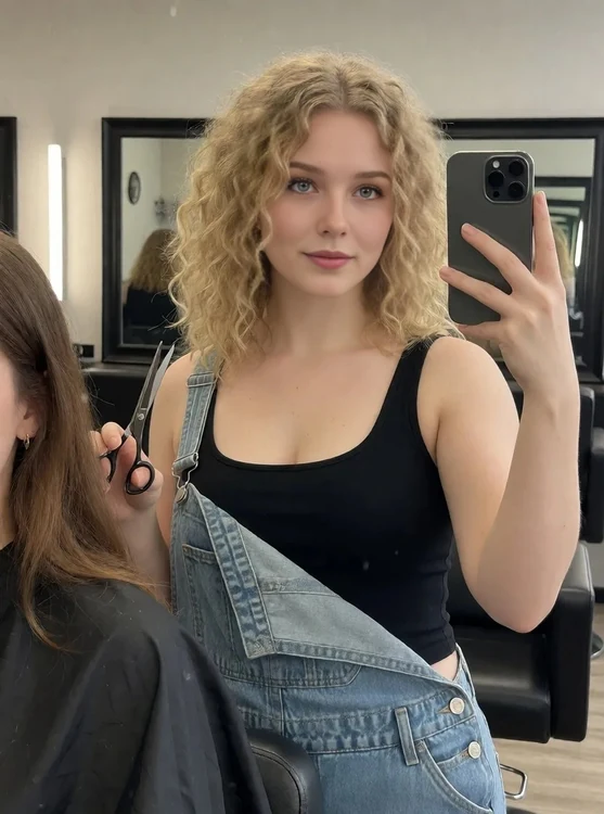 Ivy - Hair salon mirror selfie, she holds scissors near a client’s chair, denim overal